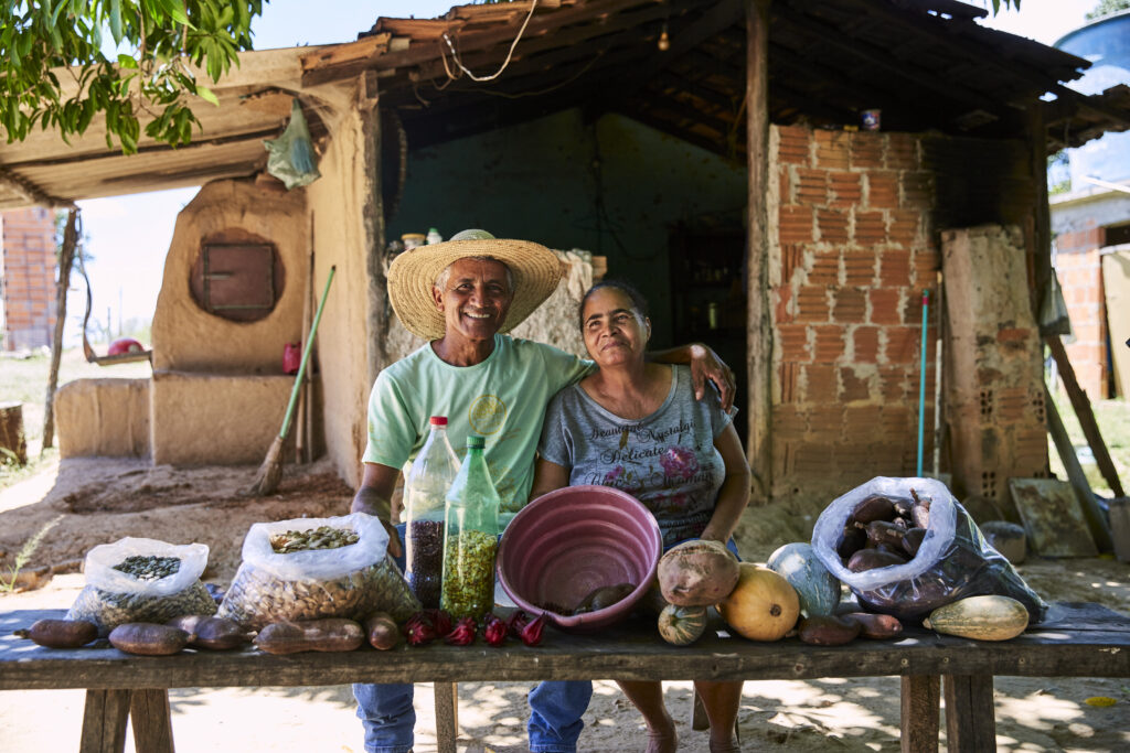 Casal de agricultores sorrindo atrás de uma mesa com frutos, sementes e produtos do Cerrado, diante de uma casa de adobe
